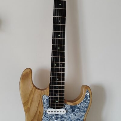 Primer plano de guitarra eléctrica artesanal con cuerpo de fresno natural visto y golpeador de perloide azul. Muestra la veta de la madera y la configuración de pastillas.