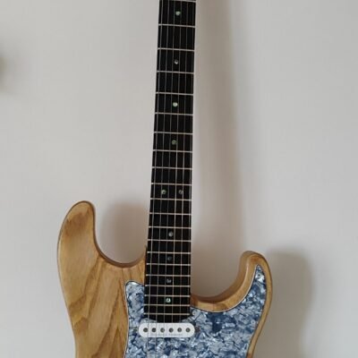 Primer plano de guitarra eléctrica artesanal con cuerpo de fresno natural visto y golpeador de perloide azul. Muestra la veta de la madera y la configuración de pastillas.