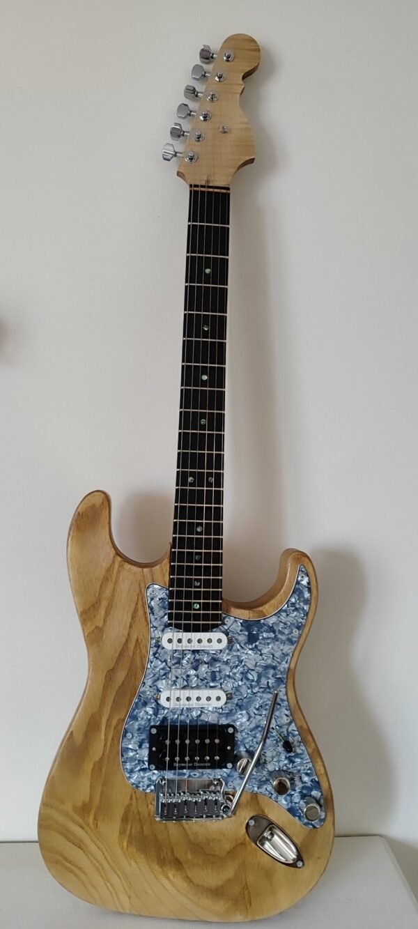 Primer plano de guitarra eléctrica artesanal con cuerpo de fresno natural visto y golpeador de perloide azul. Muestra la veta de la madera y la configuración de pastillas.